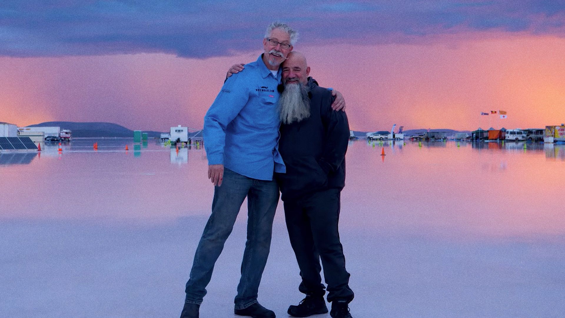 Twistgrip Tales race crew embrace on salt flats against a sunset background.