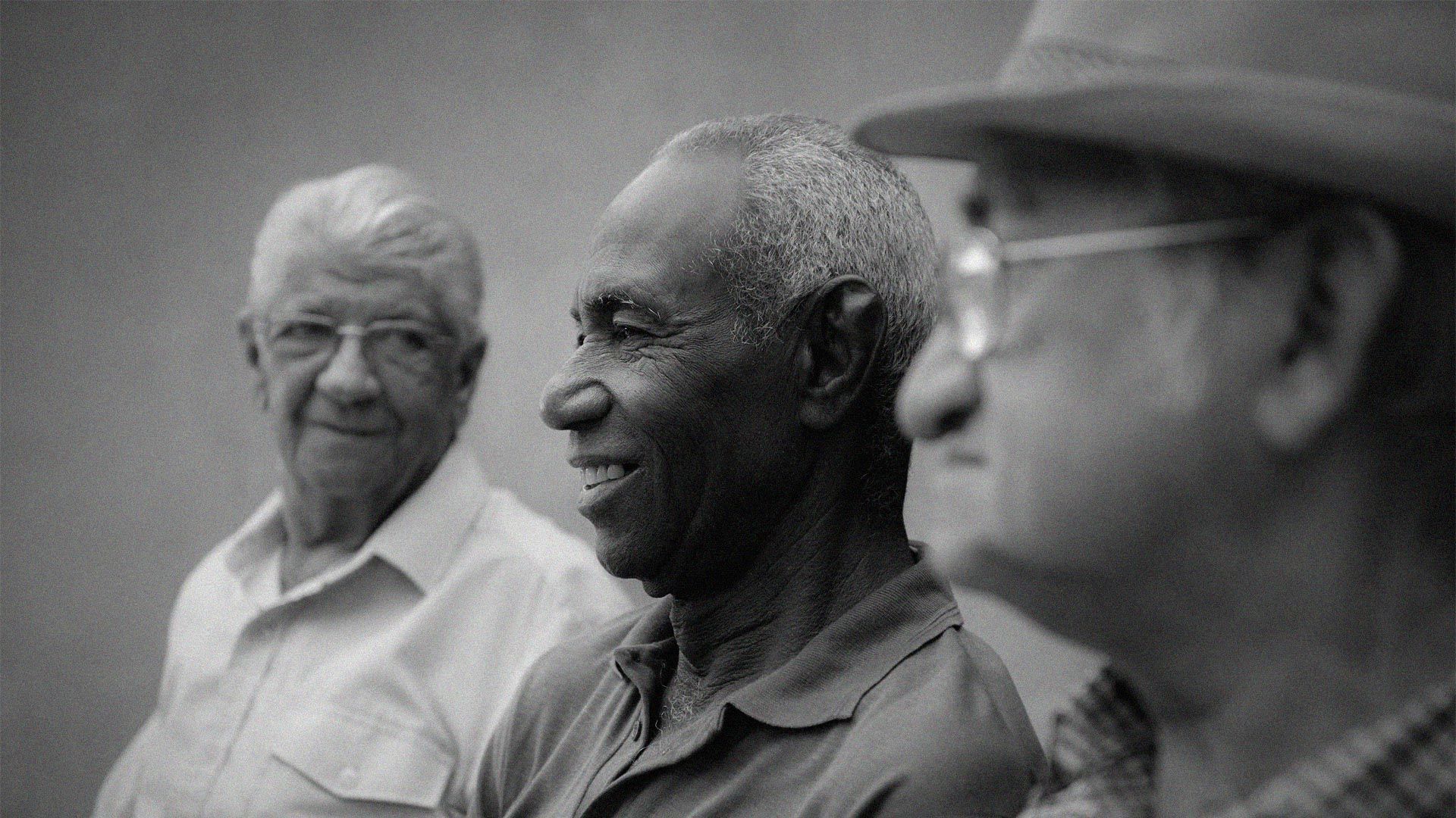 Black and white, three older men sit in a line, two looking forward and one looking back at the others