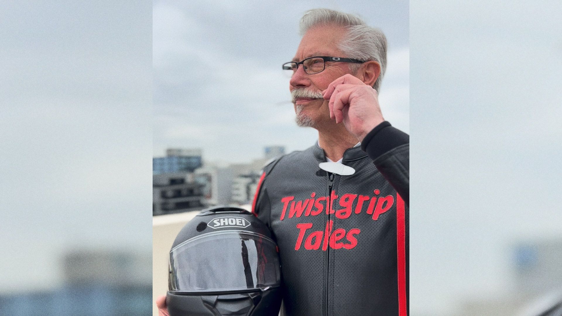 Mo Bro Bruce stands holding motorcycle helmet and twists his moustache.