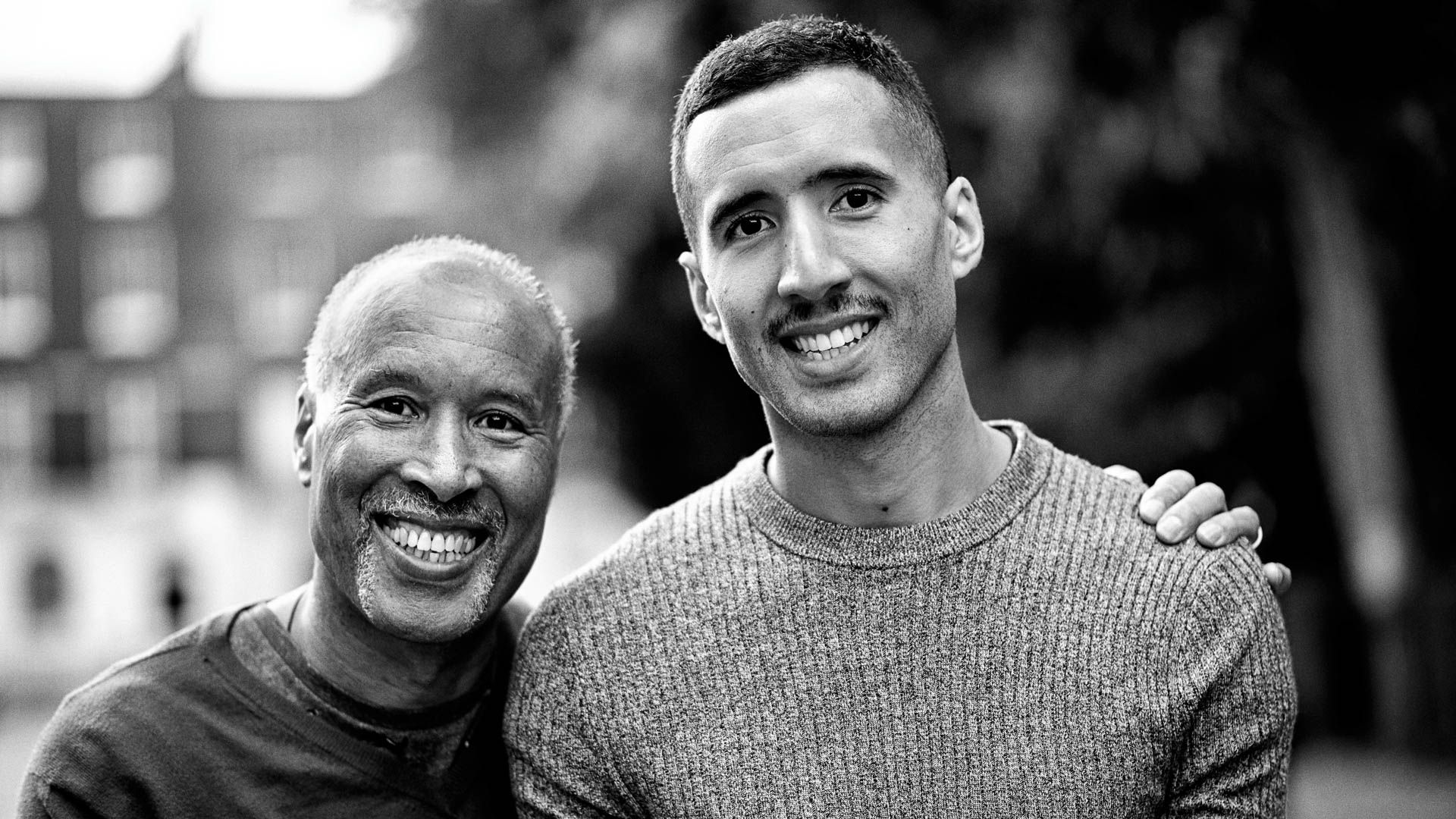 Two men smile at the camera with arm around each others shoulder, photo in black and white.