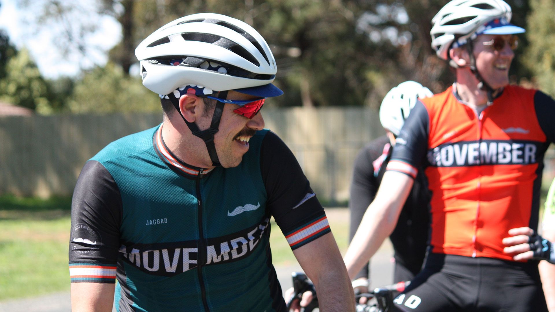 Photo of two smiling off-road bike riders, wearing Movember-branded gear and smiling on their bikes.